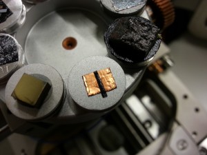 At center is an aluminum sample stub, with a square of copper tape and a strip of carbon tape. The SEM is imaging an area showing all three surfaces.